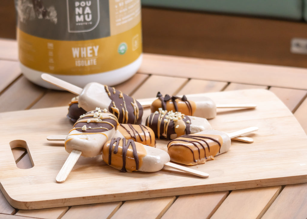 Pounamu Protein chocolate and peanut butter covered popsicles on a wooded board with a container of vanilla protein isolate in the background.