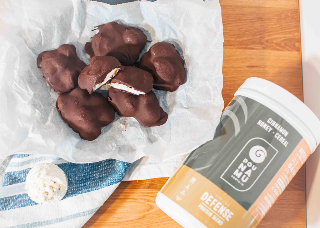 Chocolate covered protein clusters in a bowl lined with parchment paper with a scoop and container of Cinnamon, Honey and Cereal Immune Defense protein blend sitting on a wooden countertop with a blue and white striped tea towel. 
