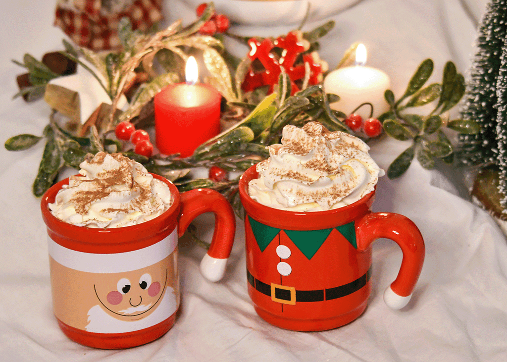 Santa and elf mug full of protein hot chocolate with whip cream on top. Candle, mini trees and wreath Christmas decorations in the background.