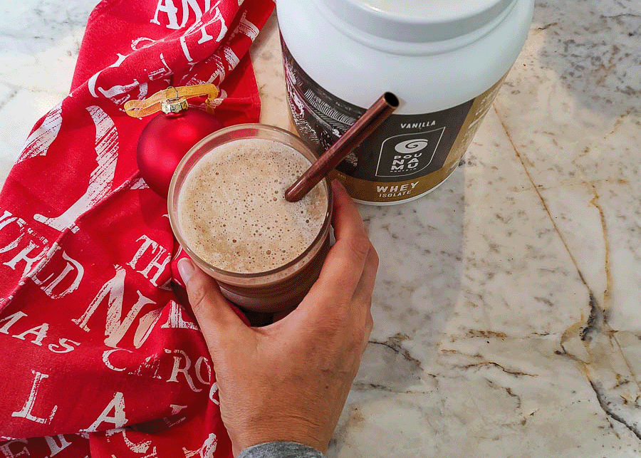 Hand holding a glass of gingerbread banana protein shake with a copper colored reusable straw which is sitting on a red Christmas tea towel, there is a red Christmas bobble and a container of Pounamu Protein Vanilla Whey Protein Isolate in the background.