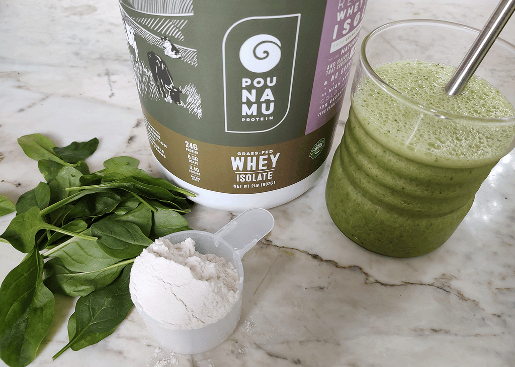 Pounamu spinach banana protein shake in a glass with a container of Berry Cream Whey Protein Isolate and a pile of fresh spinach on a marble background.